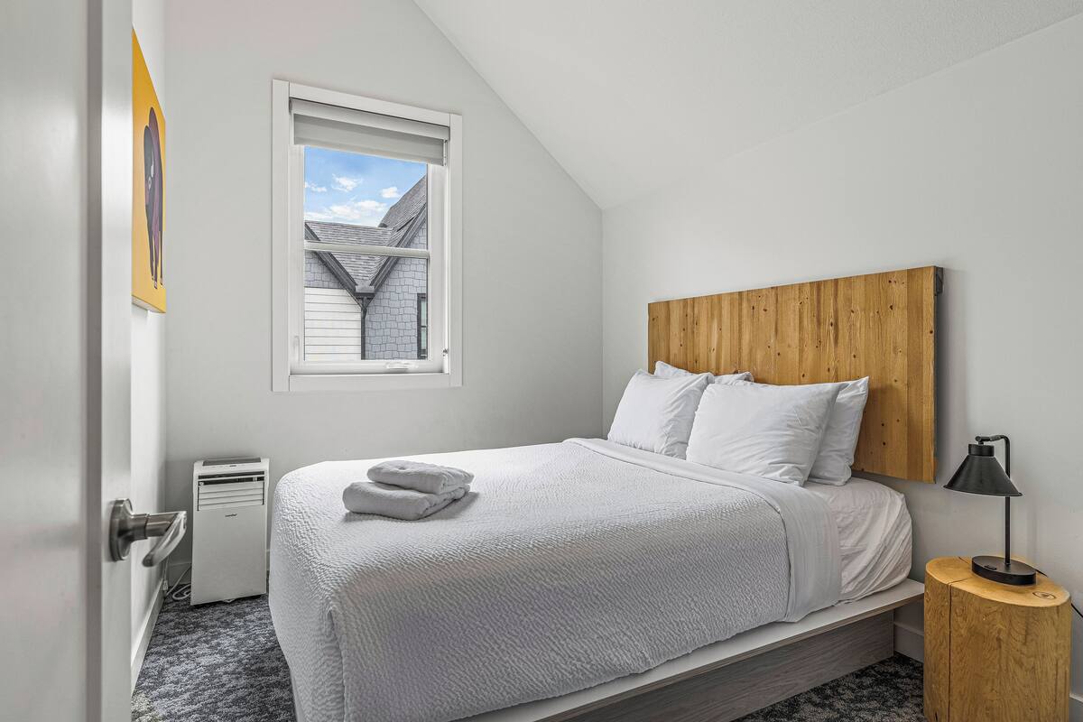 A bedroom with a bed, a wooden headboard, a bedside table with a lamp, a window with a view of a house, and a door.