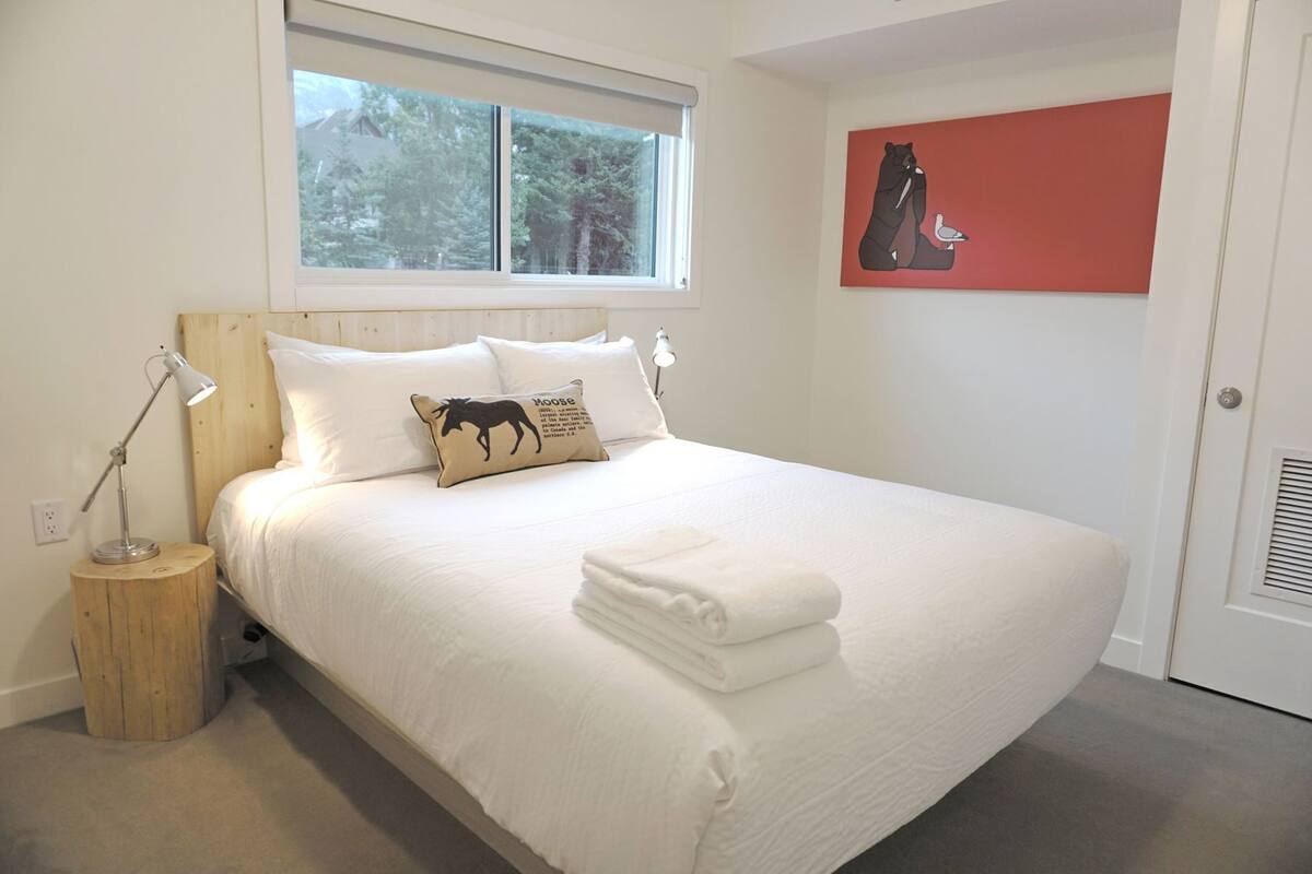 A neatly made bed with white linens, a wooden nightstand with a lamp, a window with a view of trees, and a red artwork on the wall.