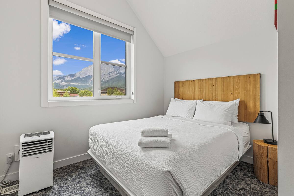A neatly made bed with white linens and a folded towel, a wooden headboard, a bedside table with a lamp, and a window with a mountain view.