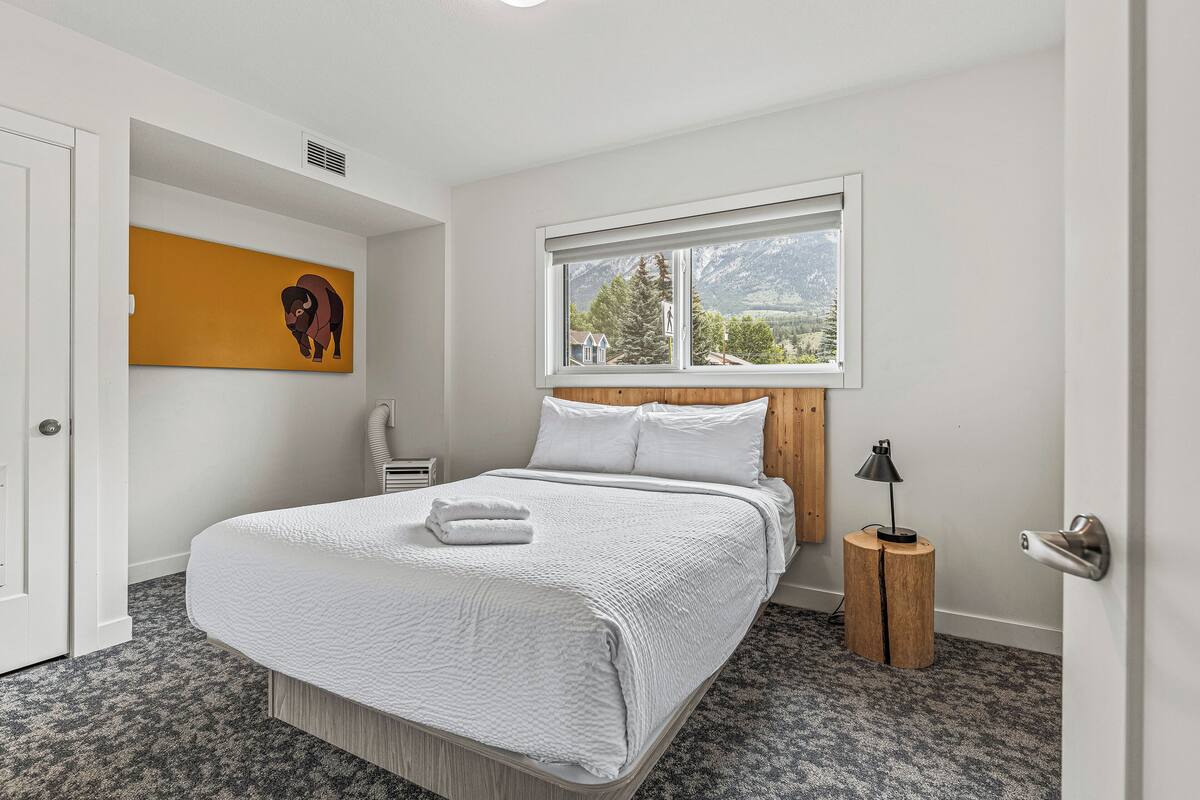 A neatly made bed with white linens, a bedside lamp, and a window with a view of mountains.