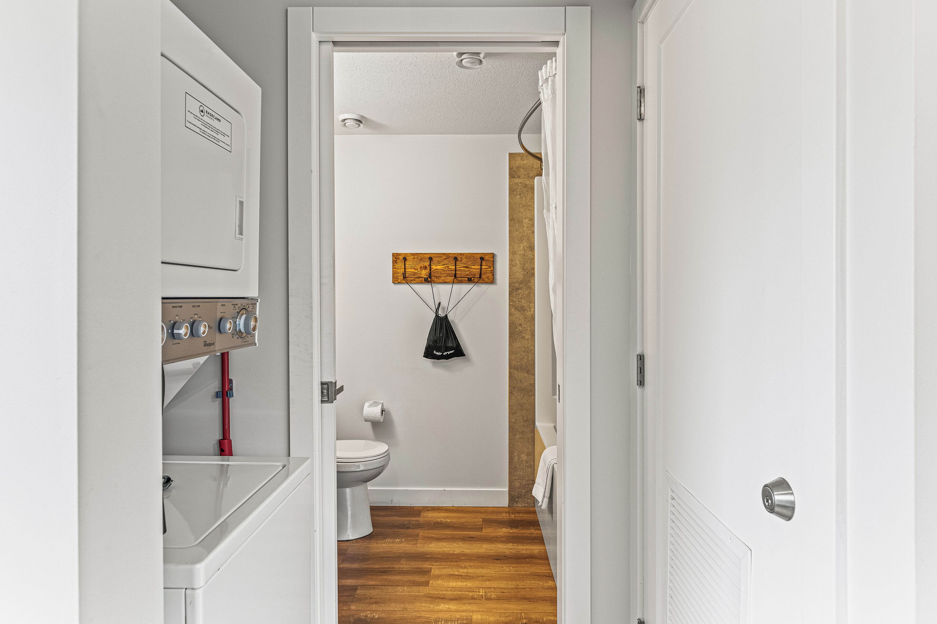 A narrow hallway with a white door on the right, a wooden floor, a white toilet, and a wall-mounted towel rack.