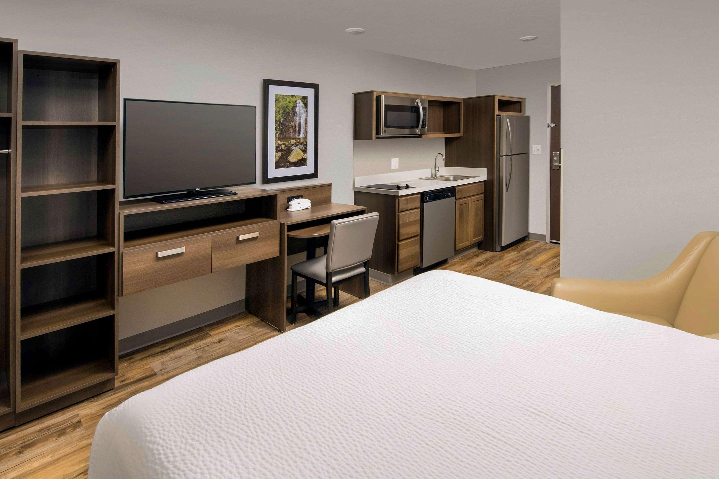 A modern hotel room with a large bed, a desk with a chair, a television, a microwave, a refrigerator, and a sink.