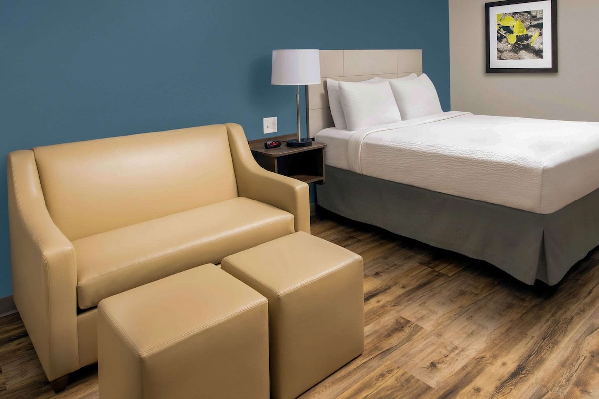 A hotel room with a beige sofa, a matching ottoman, a bed with white bedding, a nightstand with a lamp, and a framed picture on the wall.