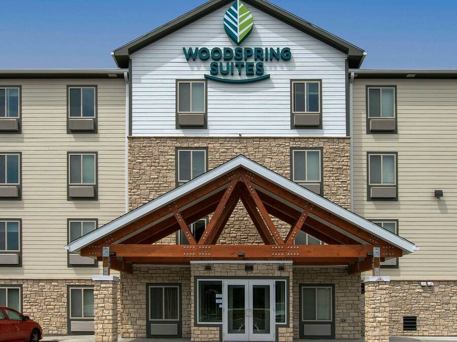 Un hotel de varios pisos con un llamativo letrero de 'Woodspring Suites', fachada de piedra y entrada cubierta.