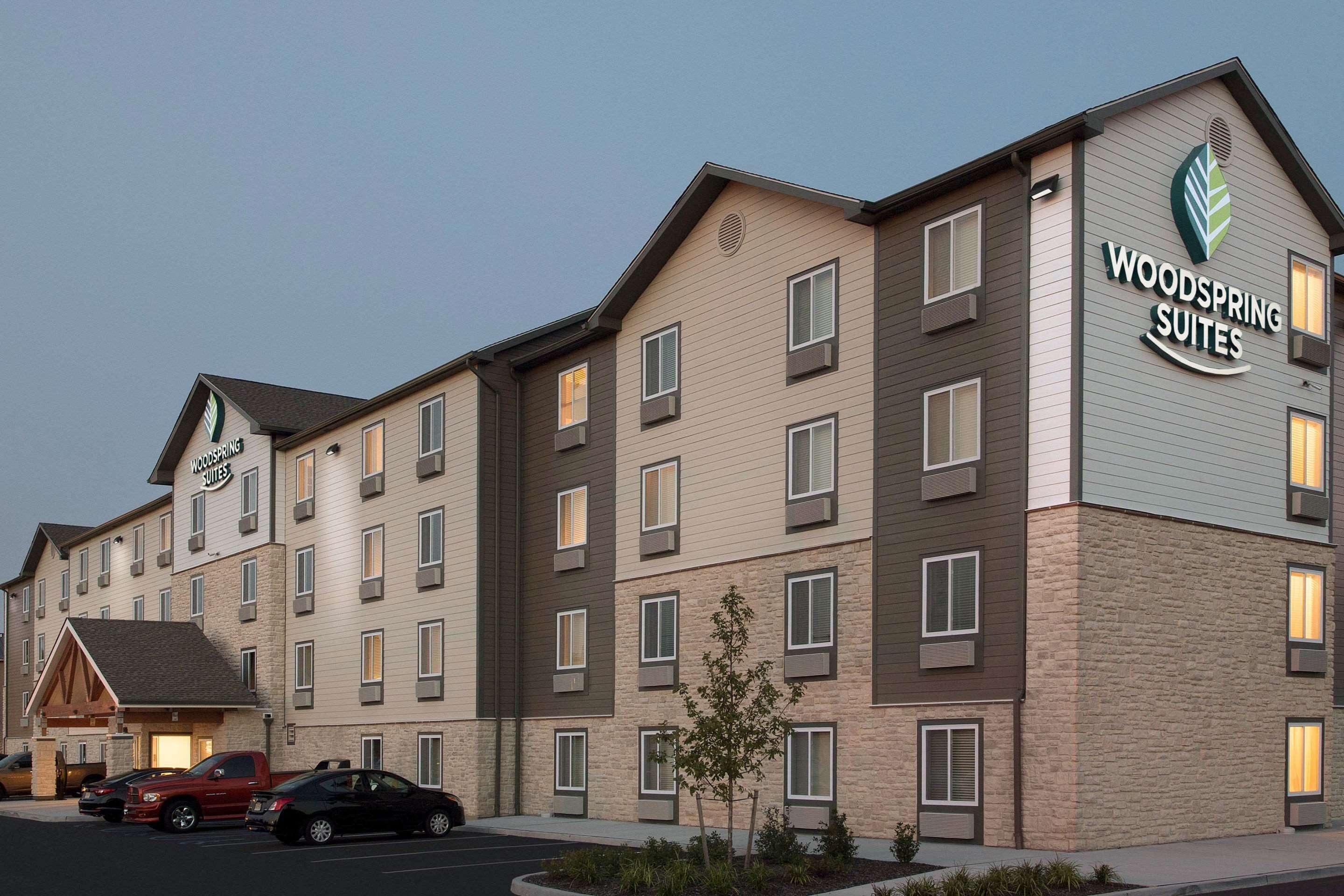 Un hotel de varios pisos con un letrero que dice "Woodspring Suites". Tiene fachada de ladrillo, múltiples ventanas y un estacionamiento con varios autos.