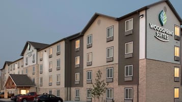 Un hotel de varios pisos con un letrero que dice "Woodspring Suites". Tiene fachada de ladrillo, múltiples ventanas y un estacionamiento con varios autos.