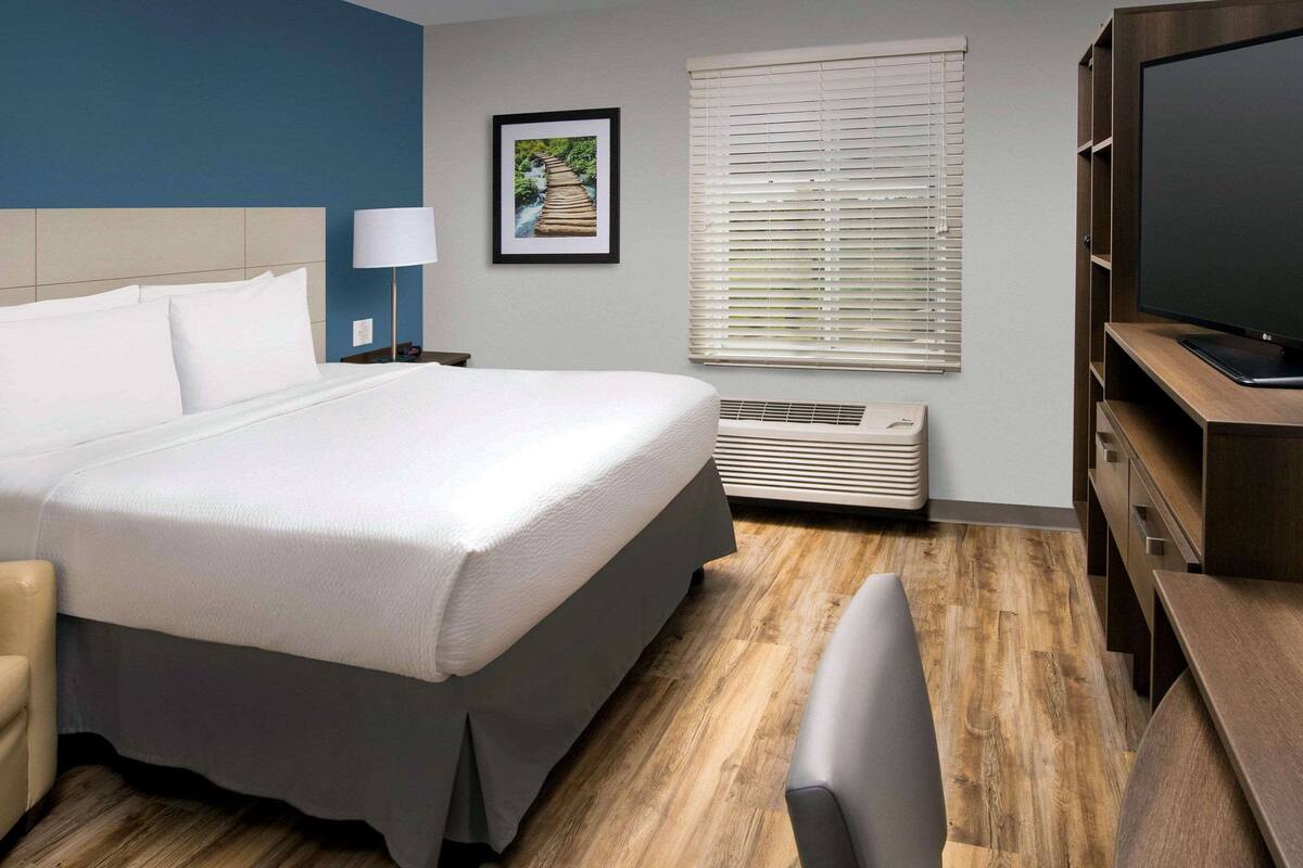 A hotel room with a large bed, a TV, a desk, a chair, a picture on the wall, and a window with blinds.