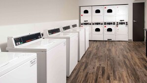 A row of white front-loading washers with control panels, a wooden floor, and a door with a handle in the background. - WoodSpring Suites South Plainfield (South Plainfield)