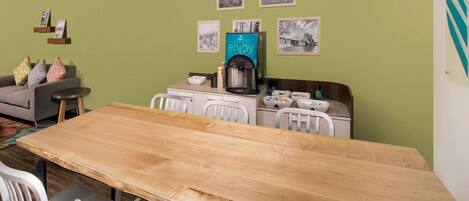 A modern kitchen with a wooden table, white chairs, a coffee machine, and a green wall with framed pictures.