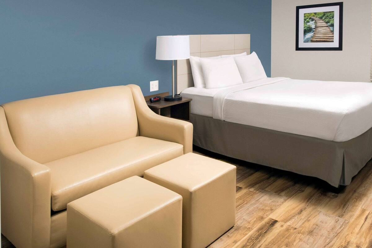 A hotel room with a beige sofa, a bed, a nightstand with a lamp, and a framed picture on the wall.
