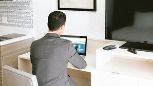 A man in a suit sitting at a desk with a laptop, a smartphone, and a pair of glasses. - WoodSpring Suites Houston Northwest (Houston)