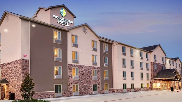 Ein mehrstöckiges Hotel mit einer Steinaußenfassade, einem gut sichtbaren Schild für WoodSpring Suites und einem gut beleuchteten Eingang.