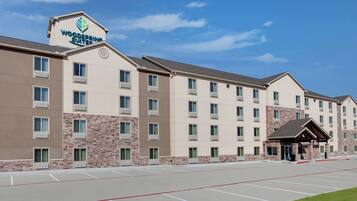 A multi-story hotel with a prominent sign reading "Woodspree Suites." It features a parking lot with marked spaces and a small grassy area in the foreground.