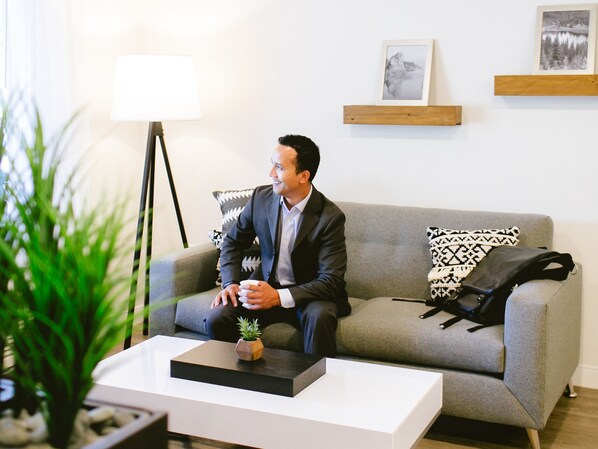 Un homme est assis sur un canapé gris dans une pièce bien éclairée, au décor moderne, avec une petite table sur laquelle se trouve une plante, et des tableaux encadrés accrochés au mur.
