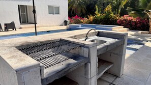 A concrete outdoor kitchen with a grill, sink, and a pool in the background.