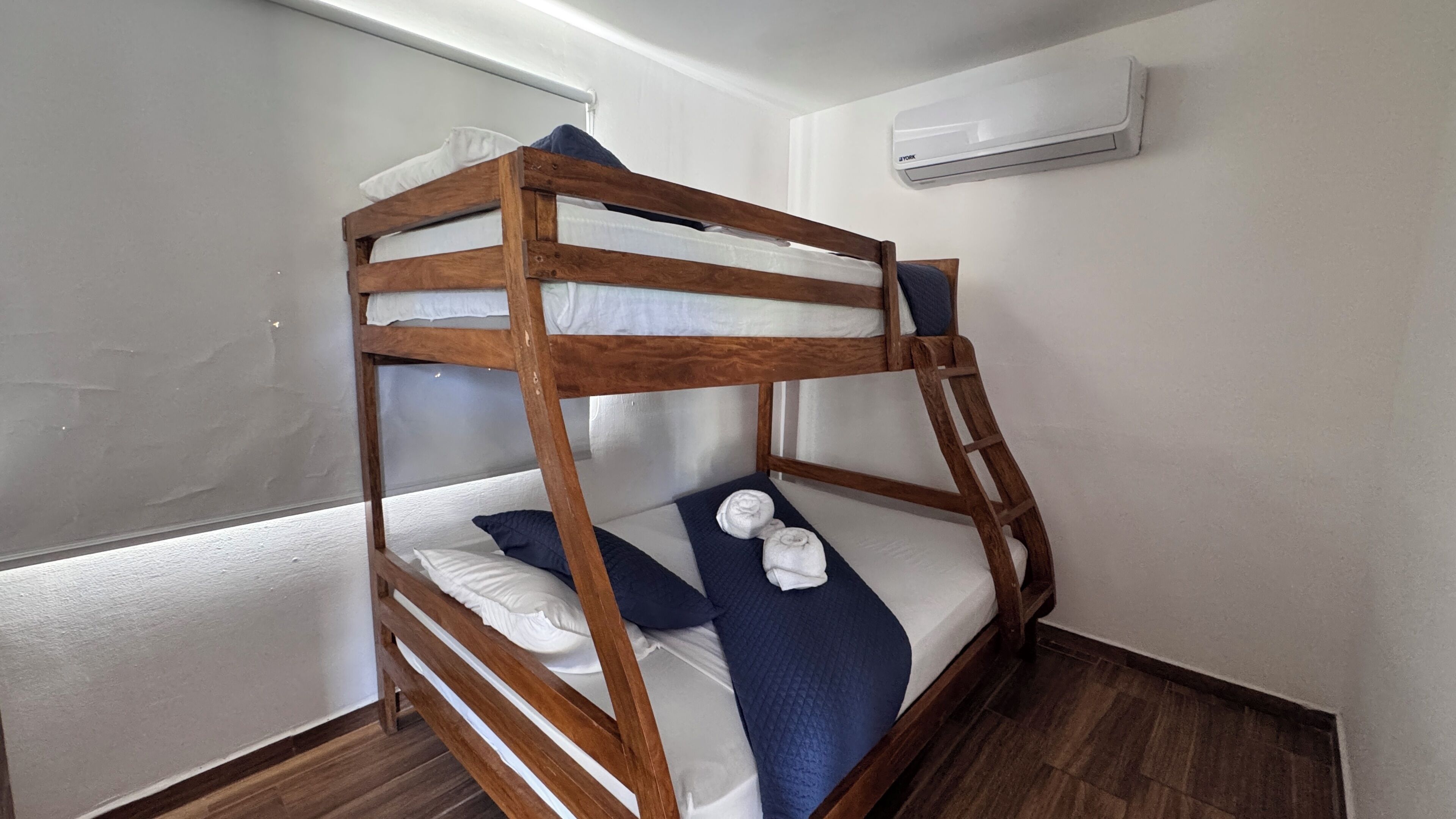 A bunk bed with white bedding and a blue blanket, wooden headboard, and an air conditioning unit on the wall.