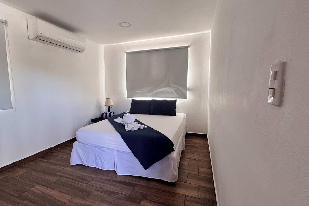 Una habitación de hotel pequeña y moderna con una cama individual, una mesita de noche con lámpara, una ventana con persianas y un aire acondicionado instalado en la pared.