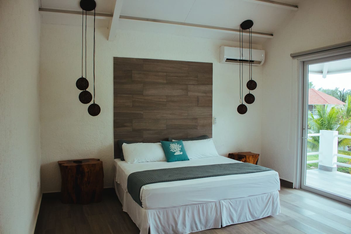 Un dormitorio moderno con cabecera de madera, cama grande con ropa de cama blanca, almohada color turquesa y lámpara colgante.
