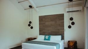Een slaapkamer met een houten hoofdbord, een bed met wit beddengoed, een blauwgroen kussen en een klein houten nachtkastje.