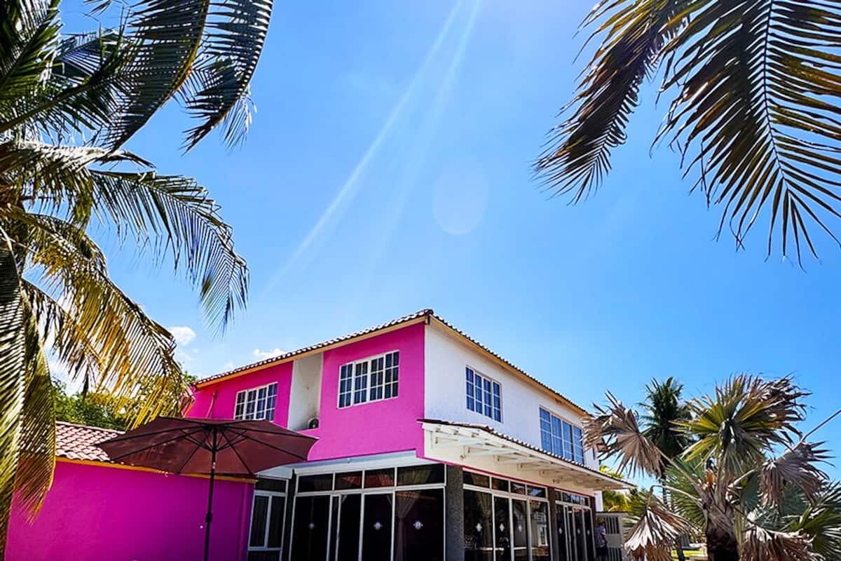 Una casa de dos pisos con piscina, rodeada de palmeras y un cielo azul despejado.