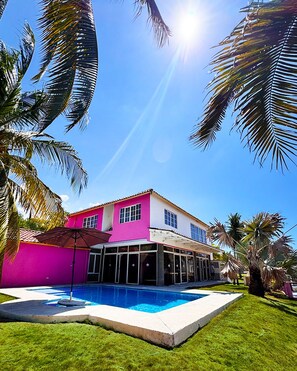 A two-story house with a swimming pool, surrounded by palm trees and a clear blue sky.