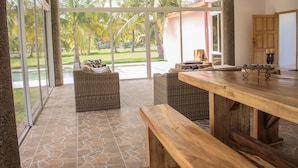Een ruime patio met rieten meubels, een houten tafel en uitzicht op palmbomen door grote glazen deuren.