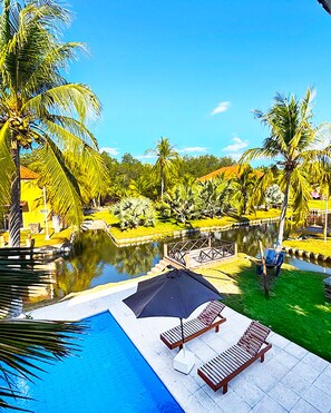 A swimming pool with lounge chairs and an umbrella, surrounded by palm trees and a small water body.