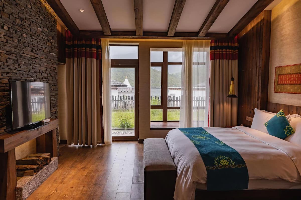 Ein Hotelzimmer mit Steinwand, Holzbalken, einem Bett mit blauer und weißer Bettdecke, einer Bank, einem Fernseher auf einem Holzregal und Blick auf einen Tempel durch eine Glastür.