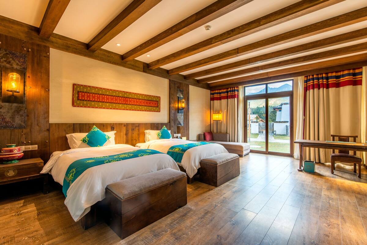 Ein Hotelzimmer mit zwei Betten, Holzbalken, einem großen Fenster mit Vorhängen und Bergblick.