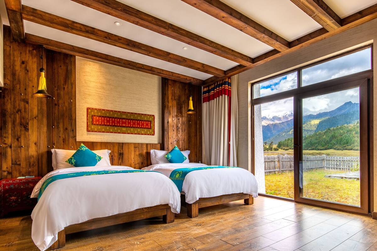 Ein Zimmer mit zwei Betten, Holzbalken, einem großen Fenster mit Blick auf die Berge und einem roten Wandbehang mit Muster.