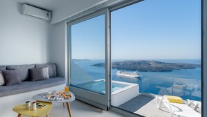 A modern living room with a large sliding glass door leading to a balcony with a view of the sea and a cruise ship. - Keti Hotel (Santorini)