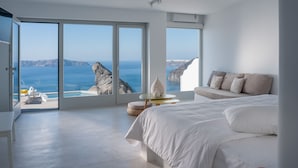 A bedroom with a large bed, a sofa, a small table, and a view of the sea and cliffs. - Keti Hotel (Santorini)