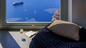 A room with a large window overlooking a coastal view, a bed with a knitted blanket, a book, and a glass of orange juice on the windowsill. - Keti Hotel (Santorini)