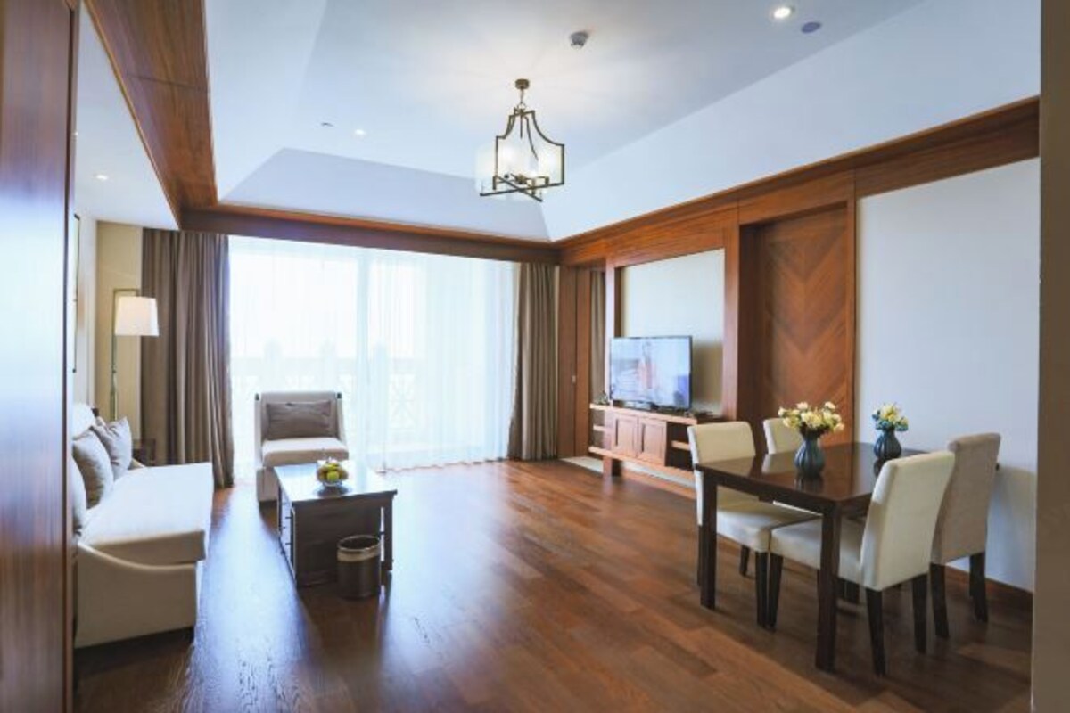 A spacious living area with a wooden floor, a sofa, a dining table with chairs, a television, and a large window with curtains.