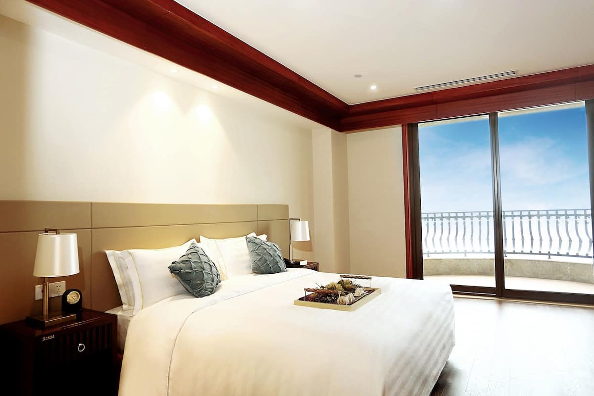 A hotel room with a large bed, a nightstand with a lamp, and a view of a balcony.