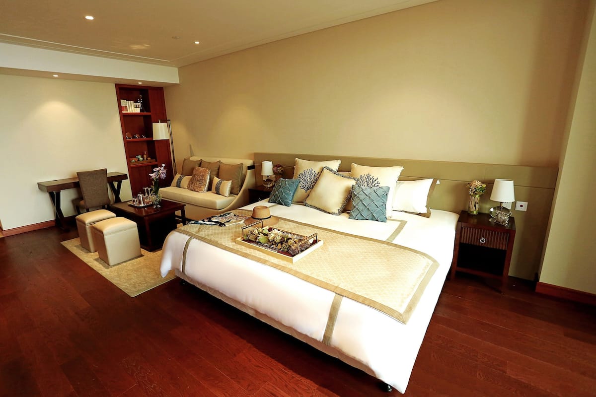 A spacious hotel room with a large bed, a seating area with a sofa and armchairs, a wooden floor, and a bookshelf.