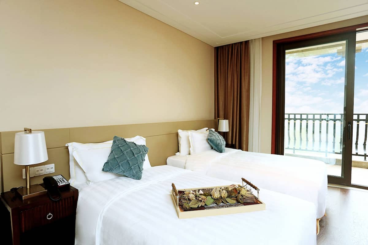 A hotel room with two beds, a nightstand with a lamp and phone, and a balcony with a view.