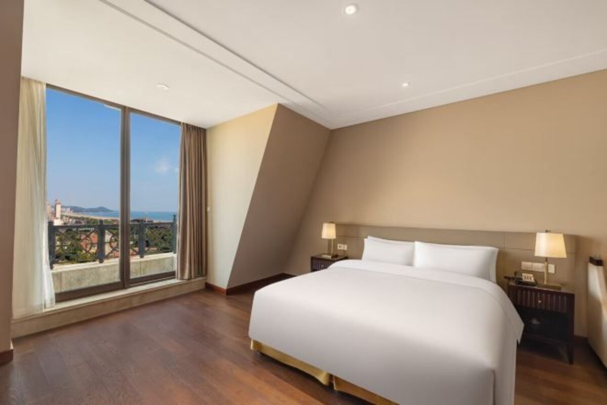 A hotel room with a large bed, two bedside tables with lamps, a view of the city through large windows, and a balcony.