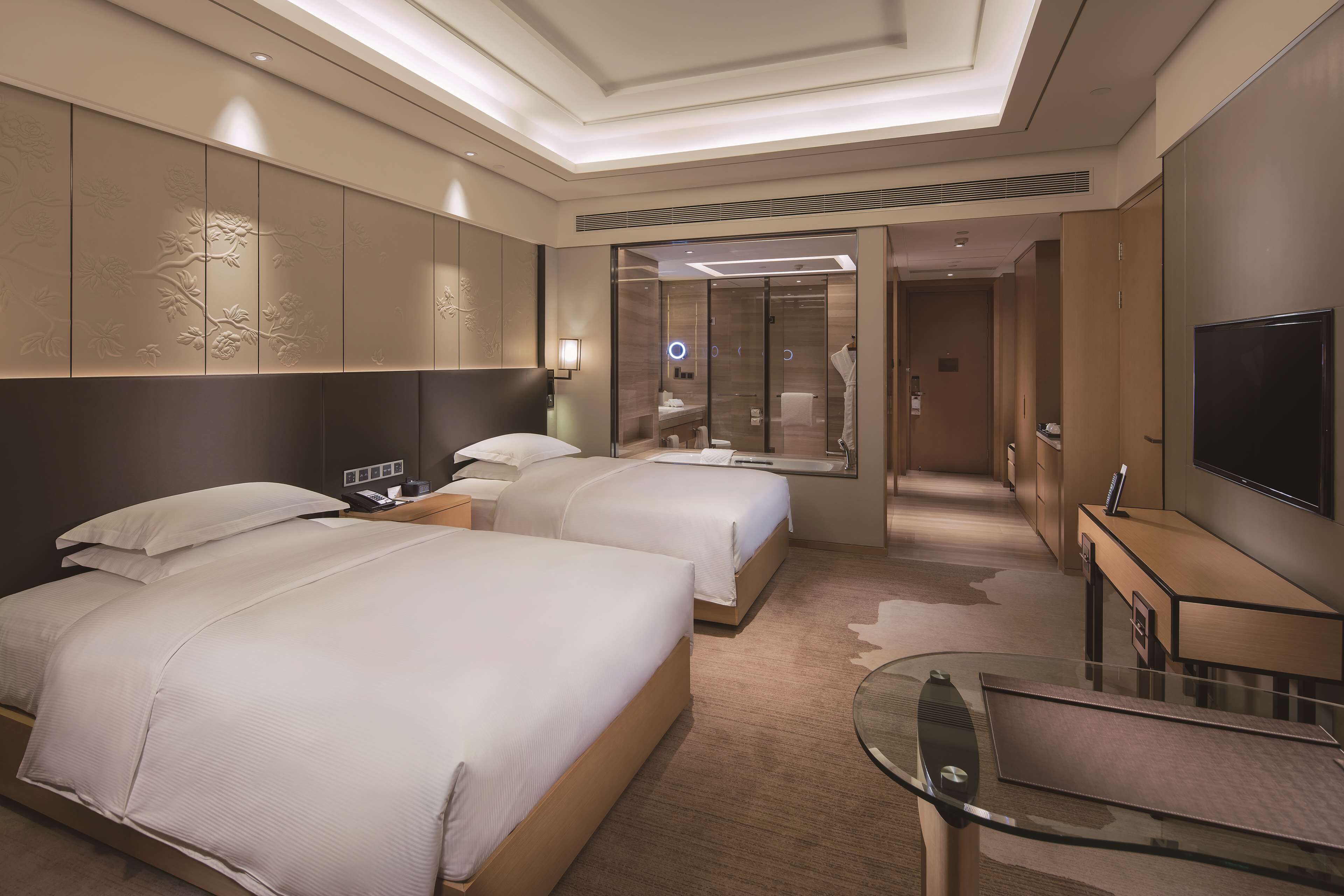 A hotel room with two beds, a TV, a glass table, and a bathroom visible through a glass door.