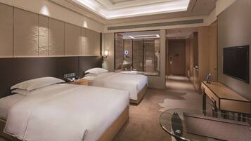 A hotel room with two beds, a TV, a glass table, and a bathroom visible through a glass door.