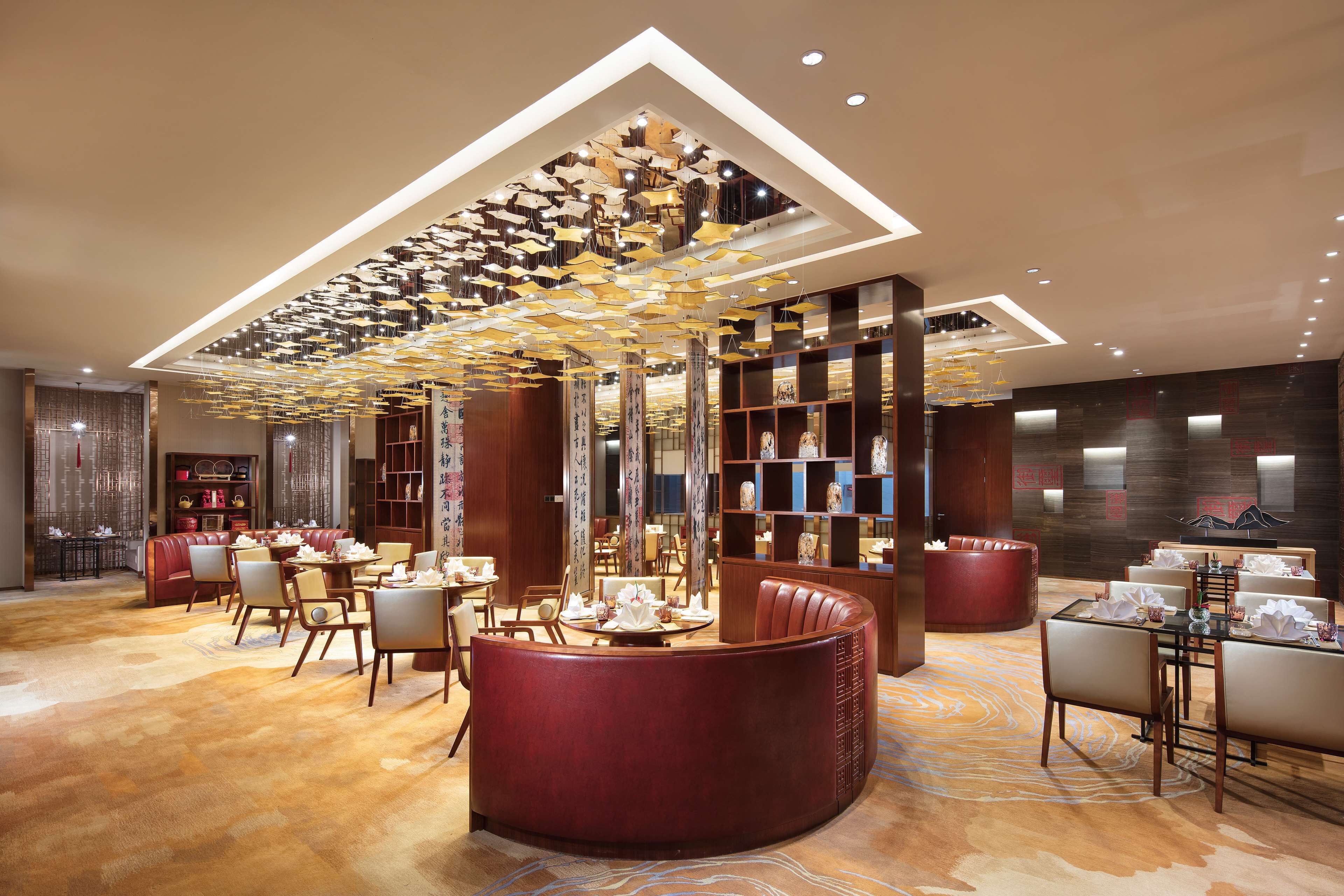Un hall d’hôtel moderne doté d’un espace salon central, d’un plafond haut avec un éclairage décoratif et de différents types de sièges.