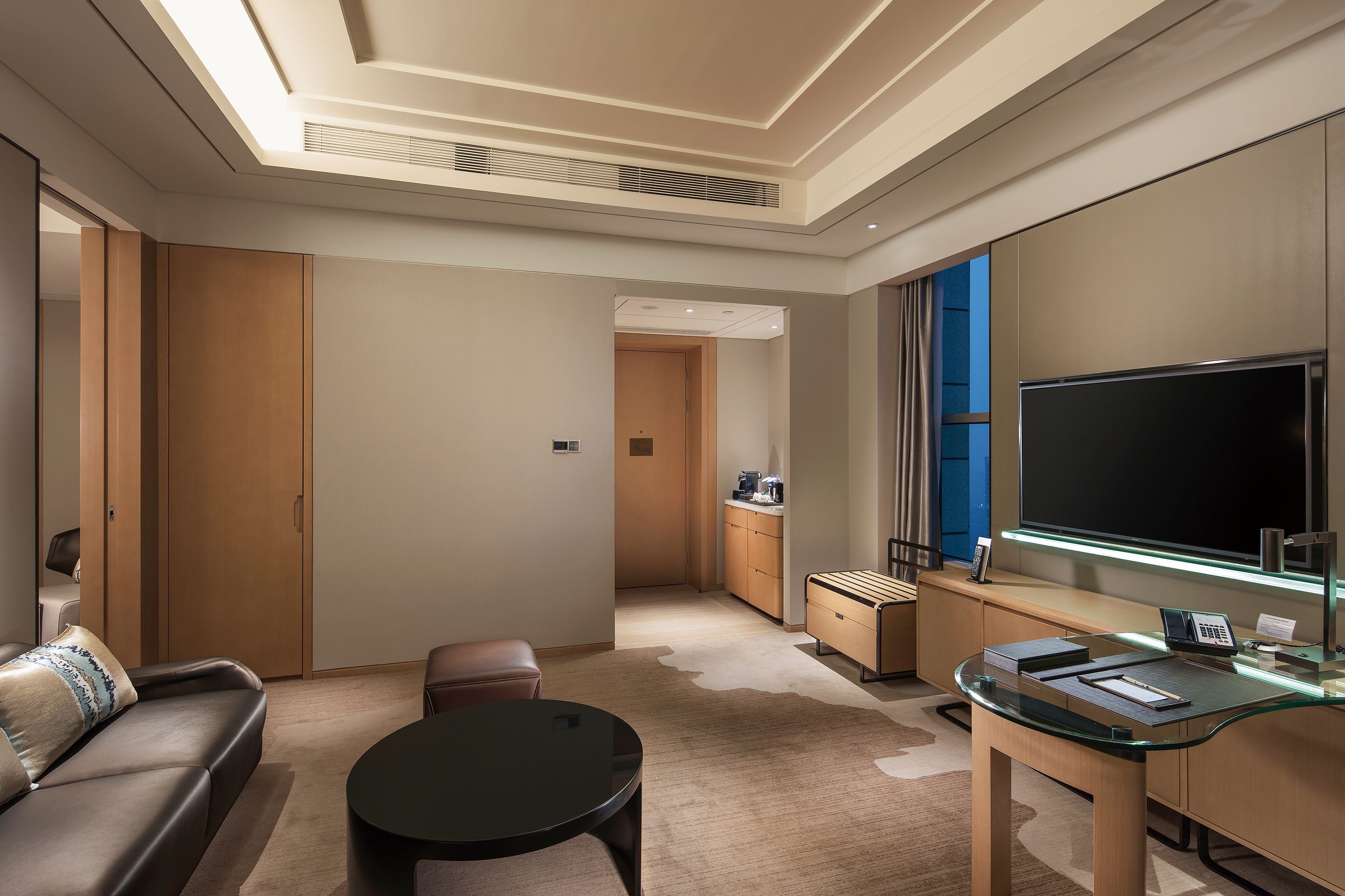 Een moderne hotelkamer met een bank, een glazen salontafel, een bureau met een flatscreen televisie en een grote spiegel.