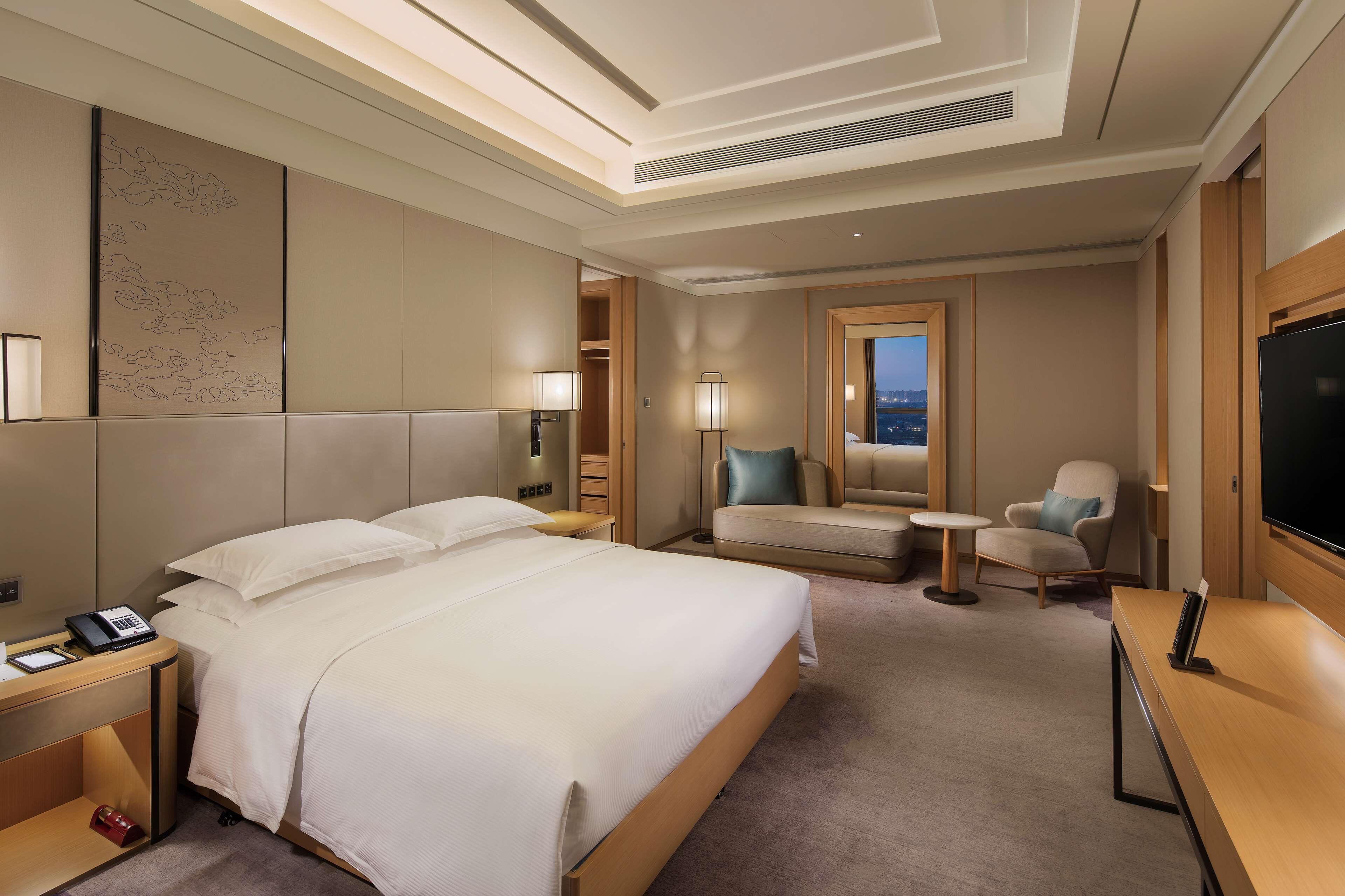 A hotel room with a large bed, a desk with a TV, a chair, a small table, and a view of the city.