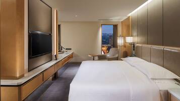 A hotel room with a large bed, a TV, a desk, a chair, and a window with a city view.