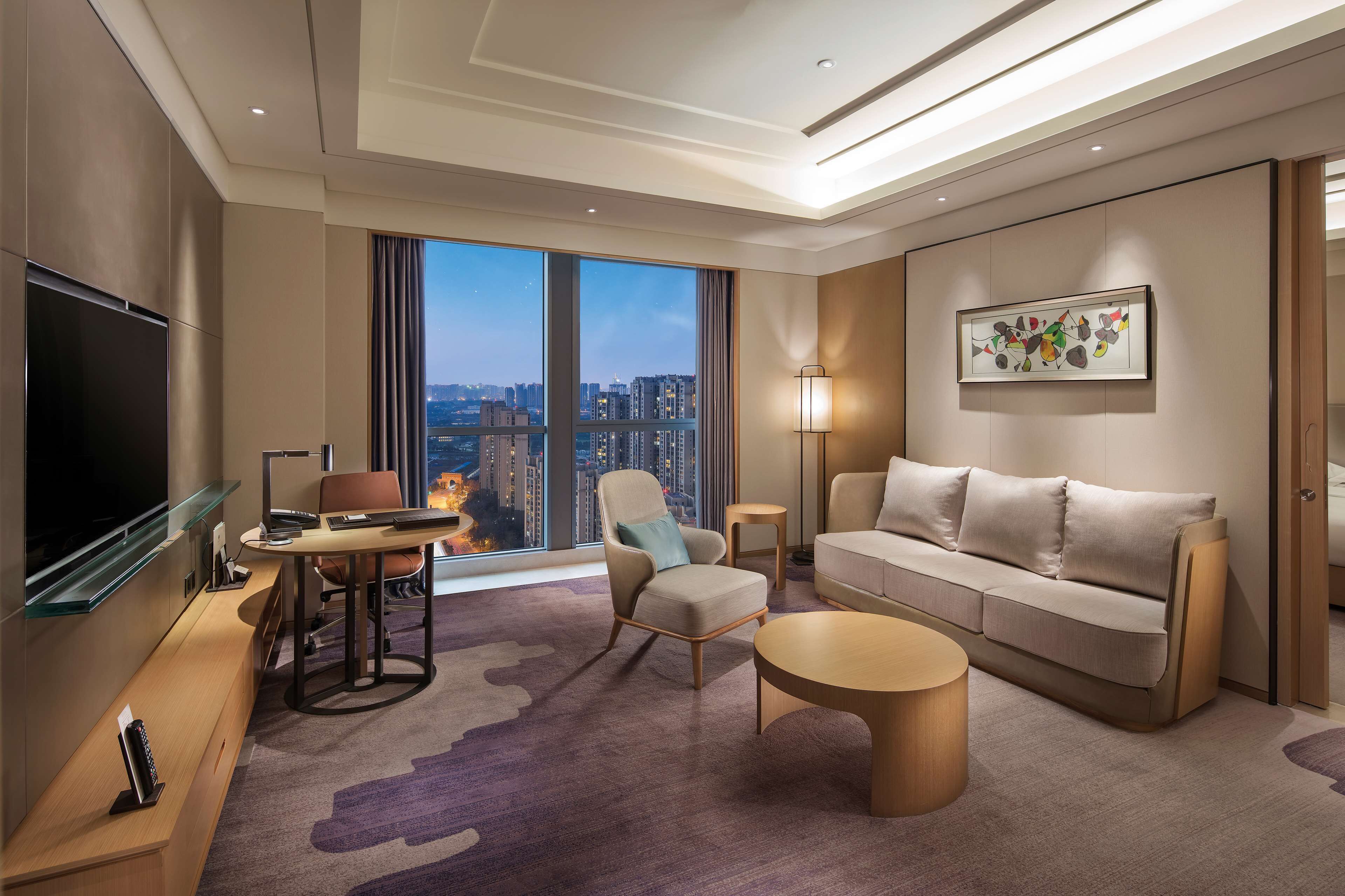 A modern hotel room with a large window offering a city view, a flat-screen TV, a sofa, a coffee table, and a desk with a chair.