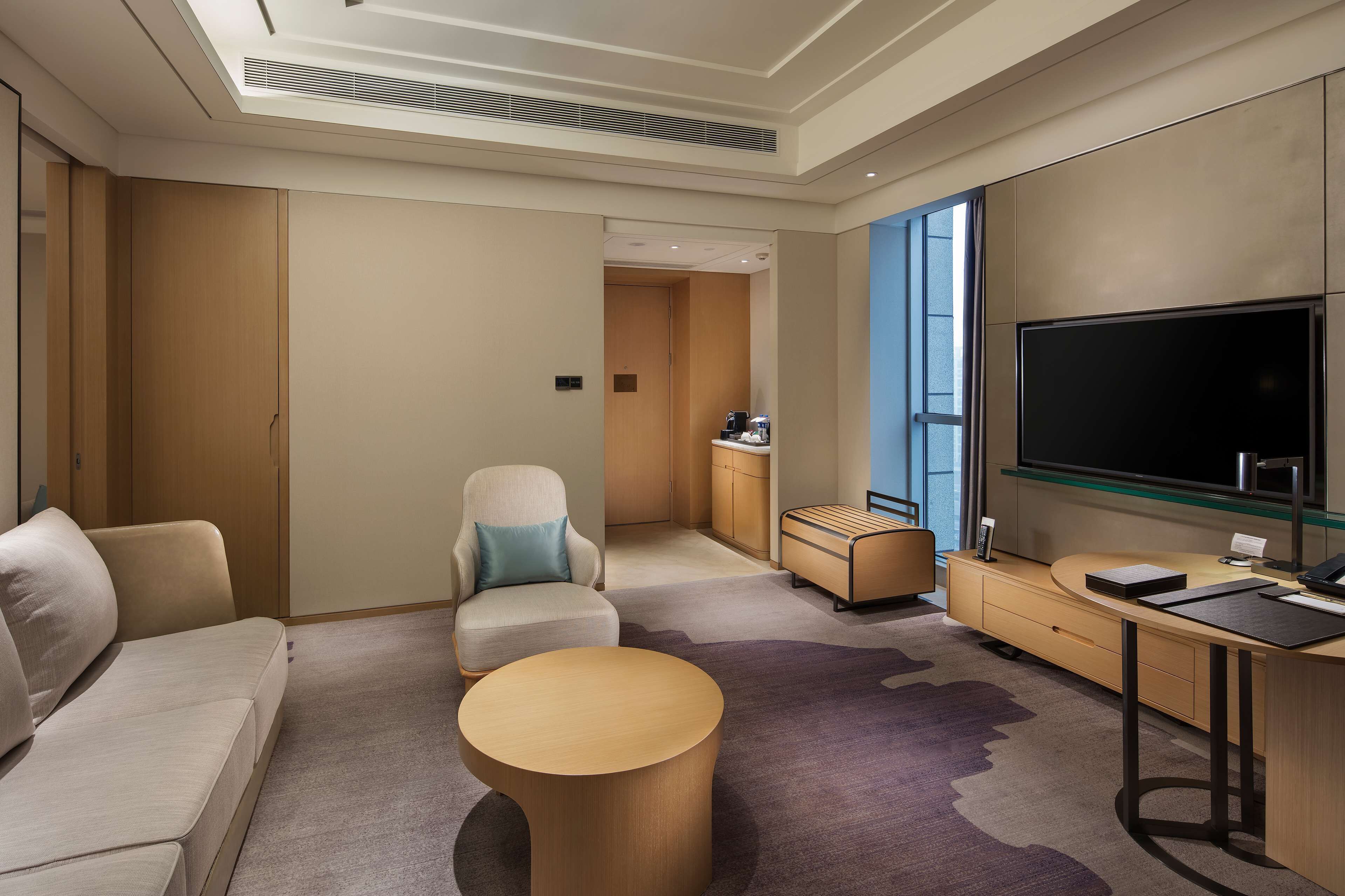 A modern hotel room with a sofa, armchair, round coffee table, TV, and a desk with a computer.