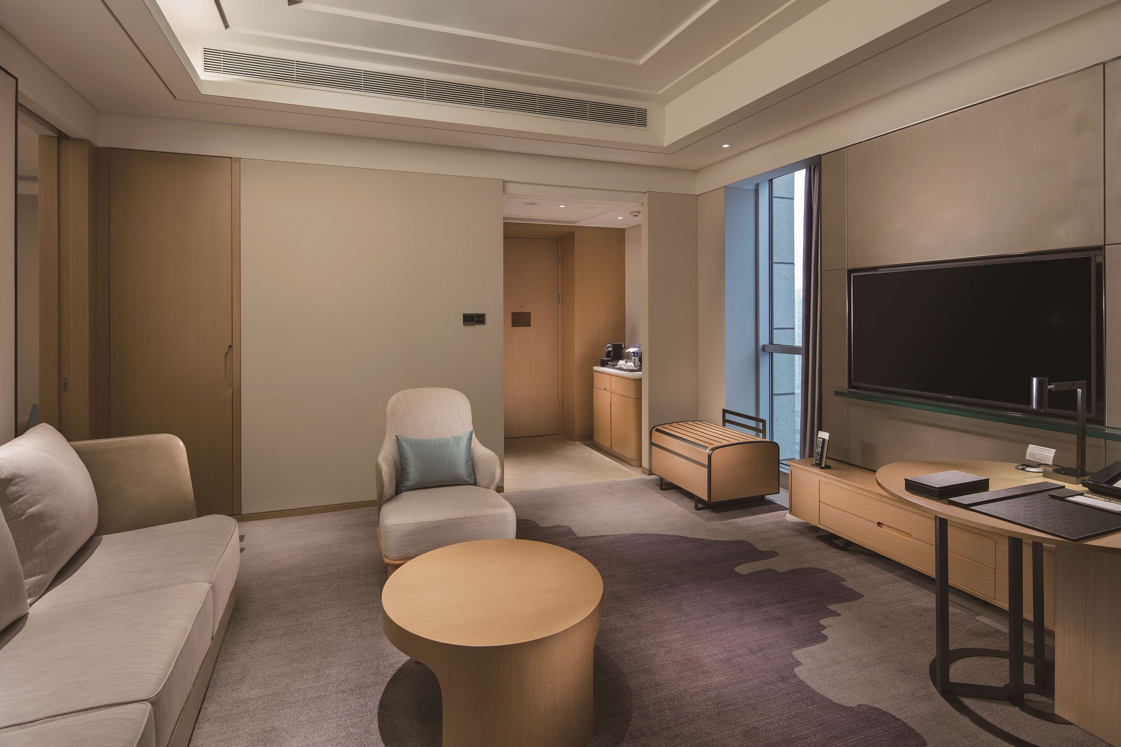 A modern hotel room with a sofa, a round coffee table, a TV, and a desk with a chair.