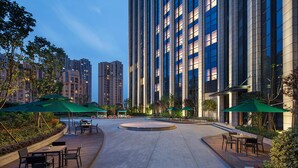 Un amplio espacio al aire libre con una zona de estar circular, rodeado de edificios altos y sombrillas verdes.