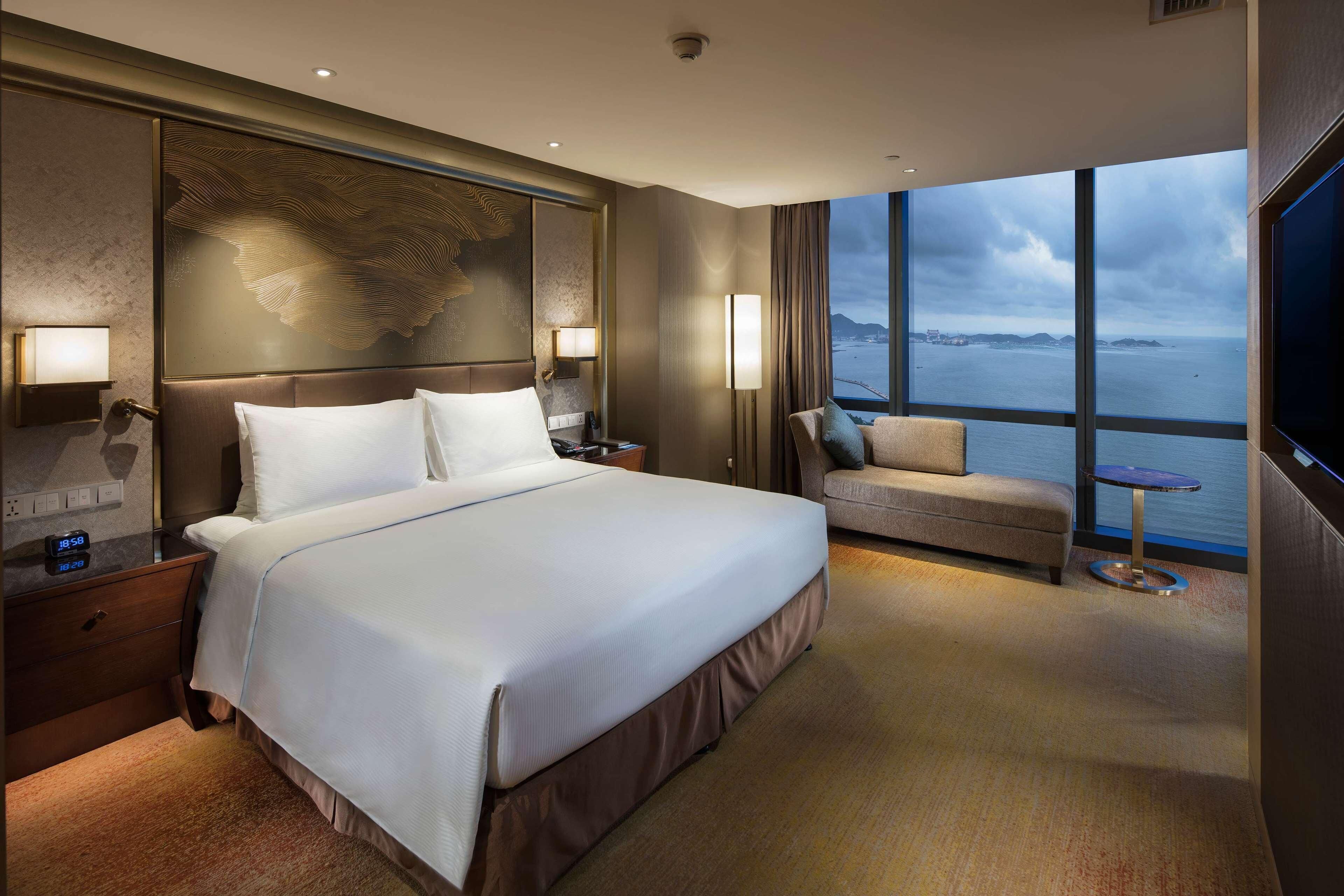 Una habitación de hotel moderna con una cama grande, un sofá, un televisor y vista al mar.