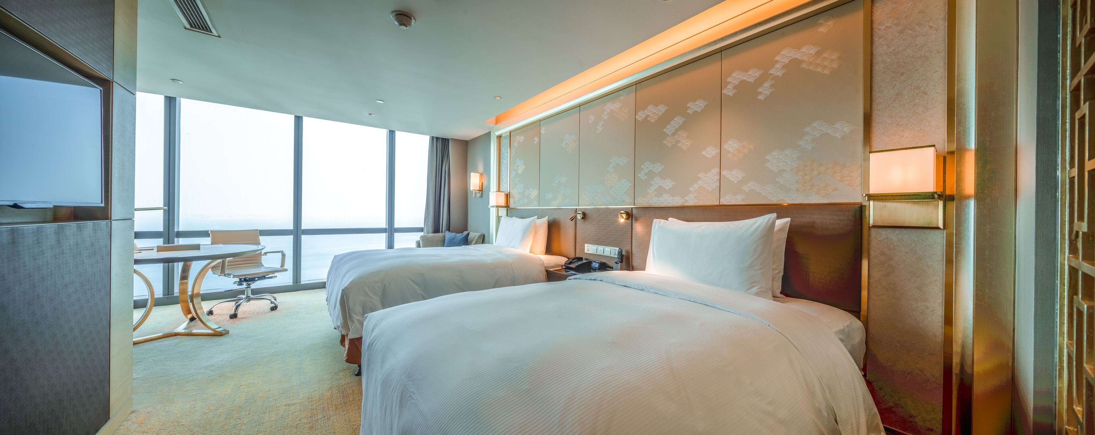 A hotel room with two beds, a desk, and a view of the city.